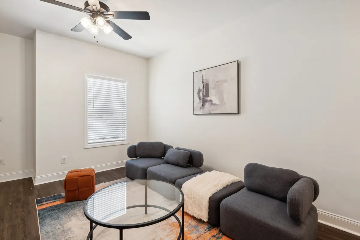 The image shows a sunlit, contemporary living room with a plush modern sofa set, a sleek glass coffee table, striking abstract wall art, and a window with blinds letting in gentle daylight. The space feels open and inviting—perfect for relaxing or spending time with friends and family.