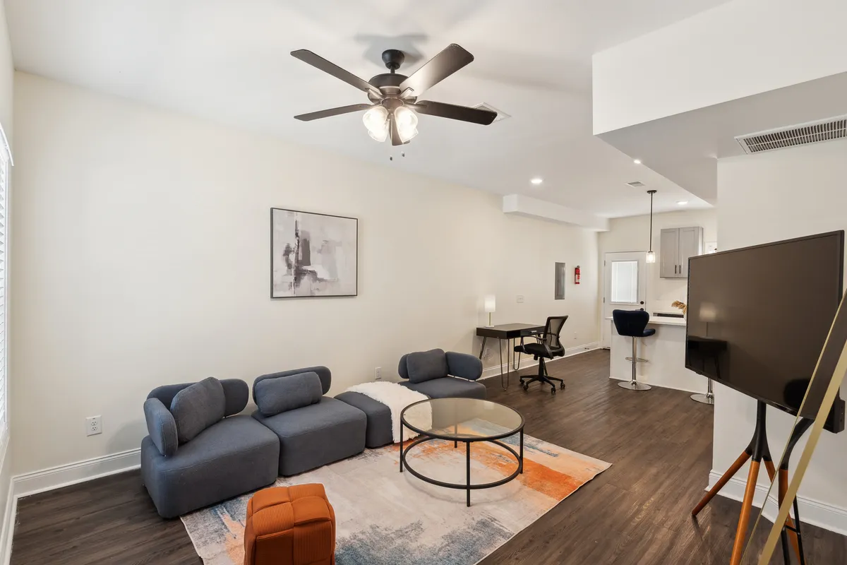 This image features a stylish, open-plan living room with a comfortable sofa, modern coffee table, wall-mounted artwork, and a television. The space is bathed in natural light, creating a welcoming environment perfect for relaxing, entertaining, or unwinding. The contemporary décor and spacious layout highlight the room’s versatility and comfort.