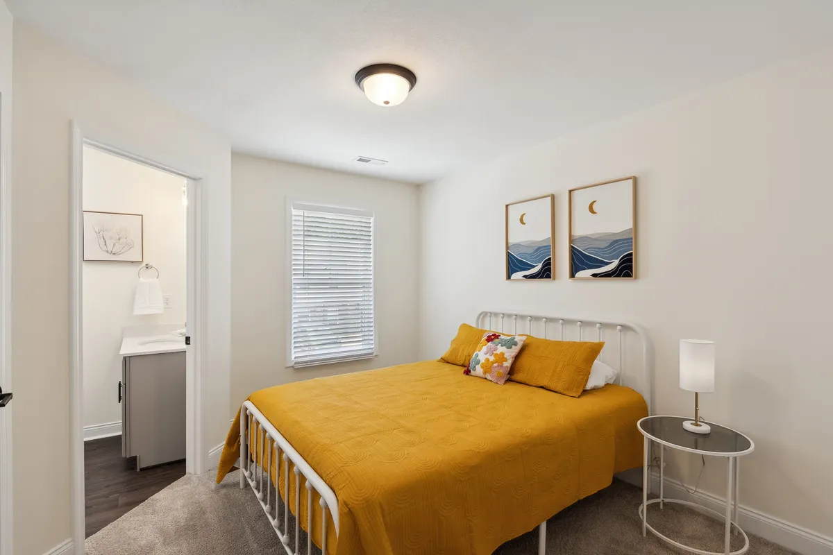 A spacious bedroom featuring a bed with a yellow quilt, vibrant artwork on the wall, and a window with blinds letting in morning light. The setting encourages relaxation, with a cozy nook by the window ideal for sipping coffee or reading a book.