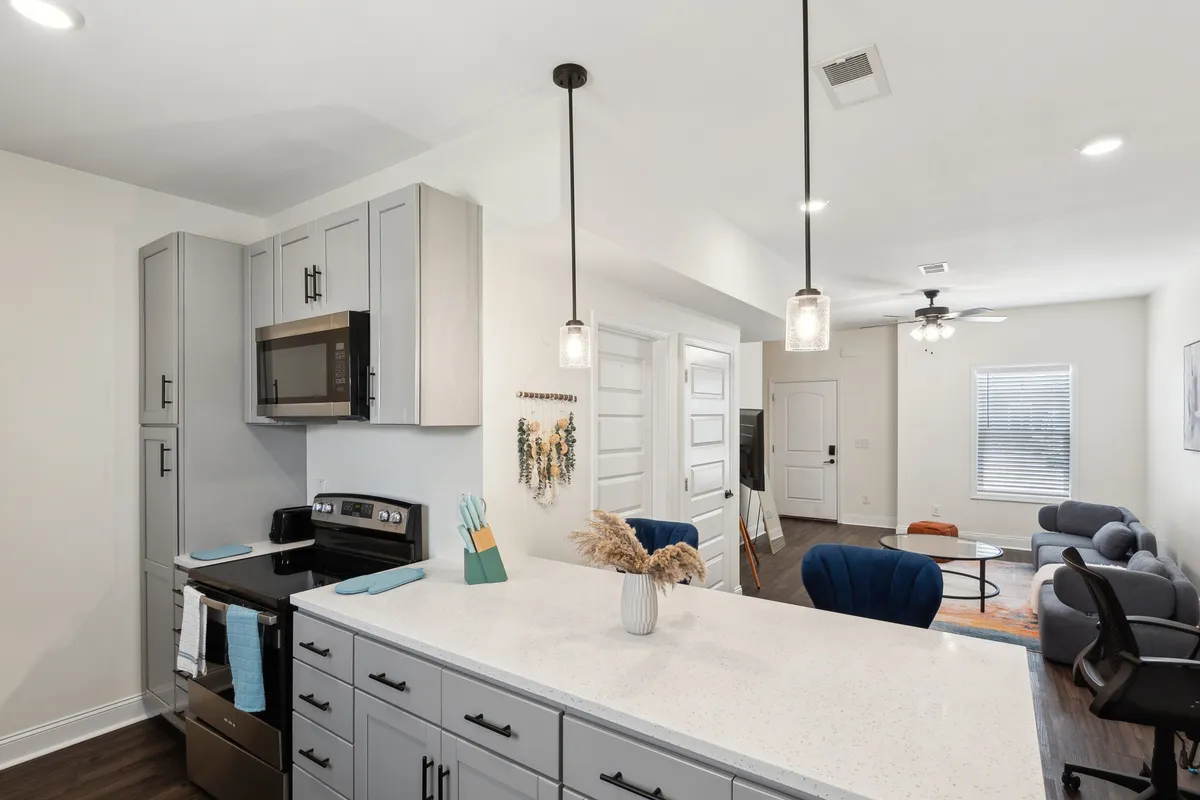 A contemporary kitchen and dining area with a prominent island, stainless steel appliances, pendant lighting, and visible living area accents. Decorative plants add a touch of warmth, and the space feels lively and ready for shared meals or entertaining guests.